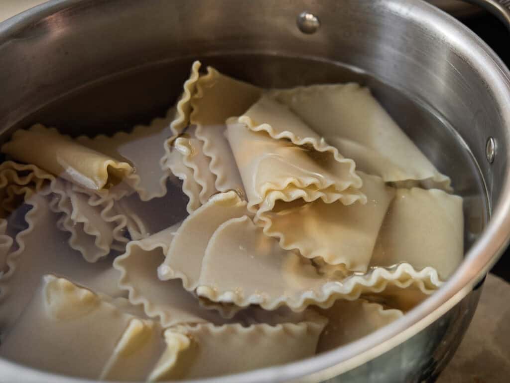 Lasagna noodles are being boiled in a stainless steel pot filled with water. The noodles are folded and partially submerged, with steam rising from the water.